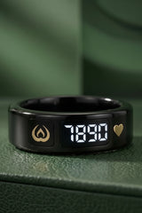 A black smart ring with a digital LED display showing '53:32', featuring gold-toned icons for flame and heart, resting on a textured green surface.